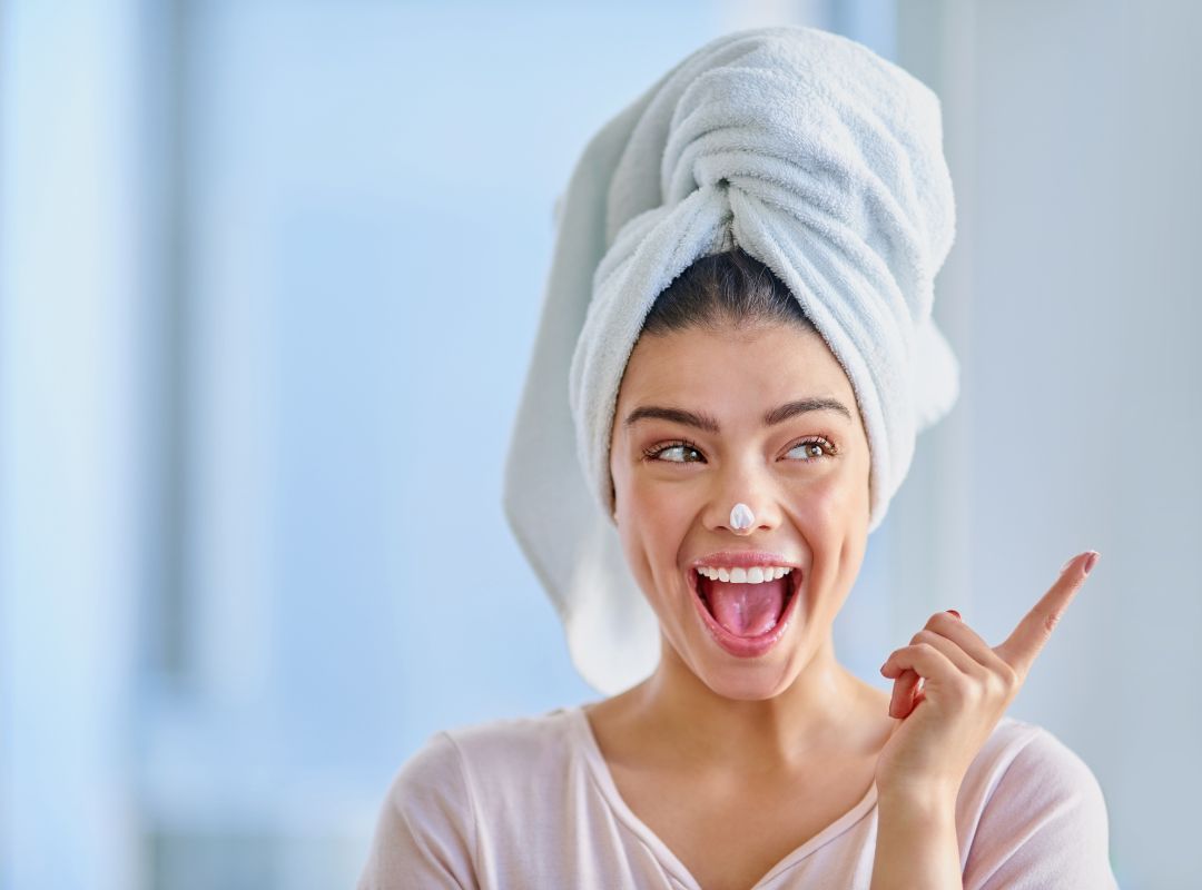 There is a woman in a light pink shirt and she has a white towel wrapped around her head. She also has moisturizer on her nose and has a happy expression and is pointing to the side. The background is a light blue and grey.