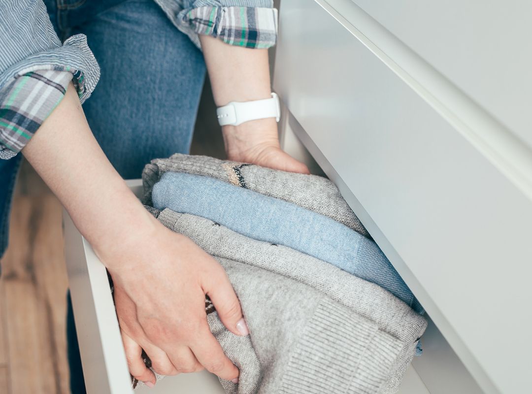 There is a person putting blue and grey sweaters in white drawers. The person has a white watch band and is wearing blue jeans and a cuffed shirt in blues, greens, and greys..