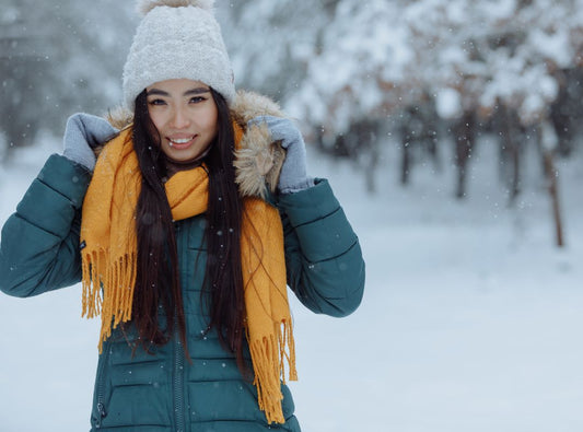 There's a woman in a white beanie, yellow scarf, and blue coat outside in the snow. There are trees behind her.