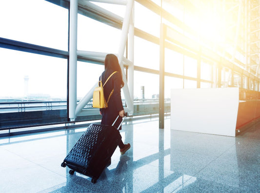 There is a woman in an airport pulling a black bag behind her and she has a yellow back pack.